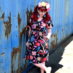 Woman dressed in a black dress with large, bold floral designs in reds, blues, and oranges, styled with a flower crown and leaning casually against a weathered blue wall. Designed by Australian designer brand Streborclothing.
