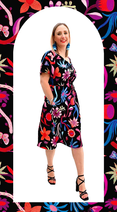 Woman dressed in a black dress with large, bold floral designs in reds, blues, and oranges, styled with a flower crown and leaning casually against a weathered blue wall. Designed by Australian designer brand Streborclothing.