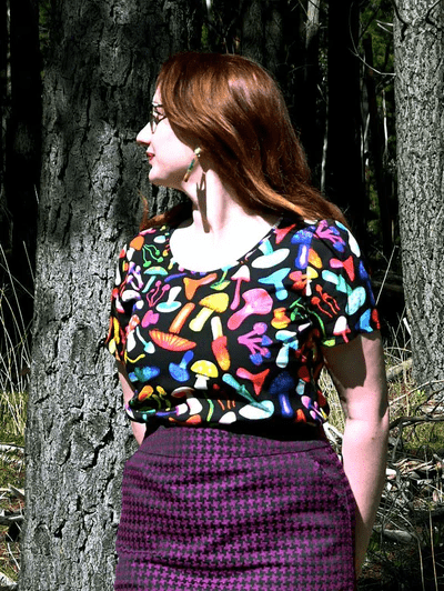 The dress features a vibrant mushroom pattern with various brightly colored mushrooms in red, yellow, blue, pink, and green on a black background. This playful design adds a whimsical and organic feel to the outfit. Designed by Australian designer brand Streborclothing.